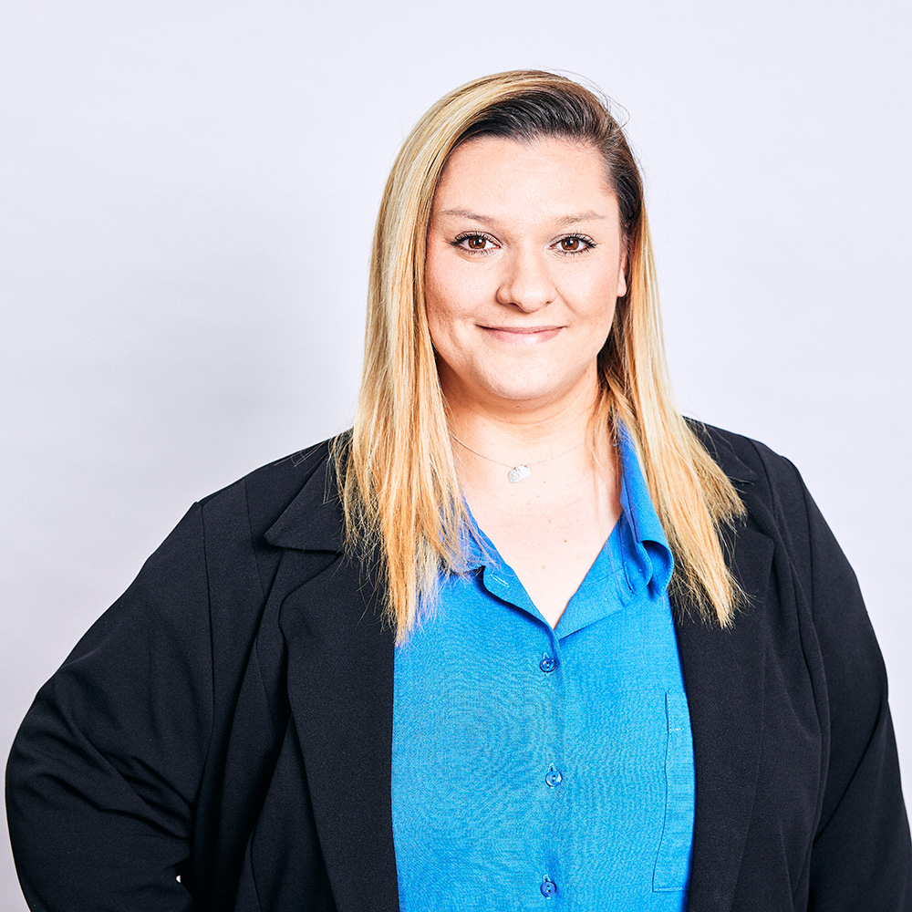 A headshot of a lady with blonde hair, wearing a blue shirt and black jacket, smiling at the camera.