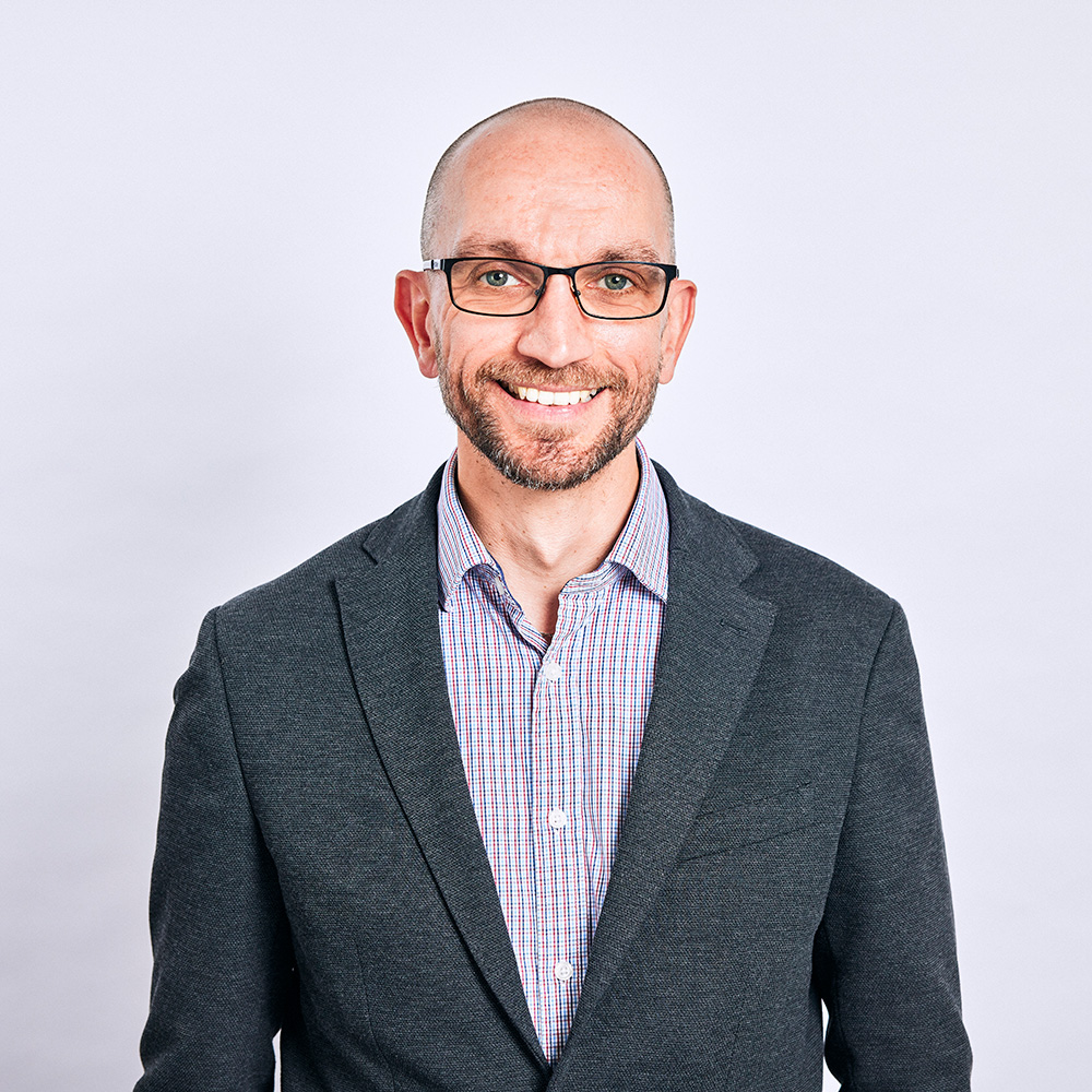A headshot of a bald man with a beard and glasses smiling at the camera, dressed in a blazer and striped shirt.