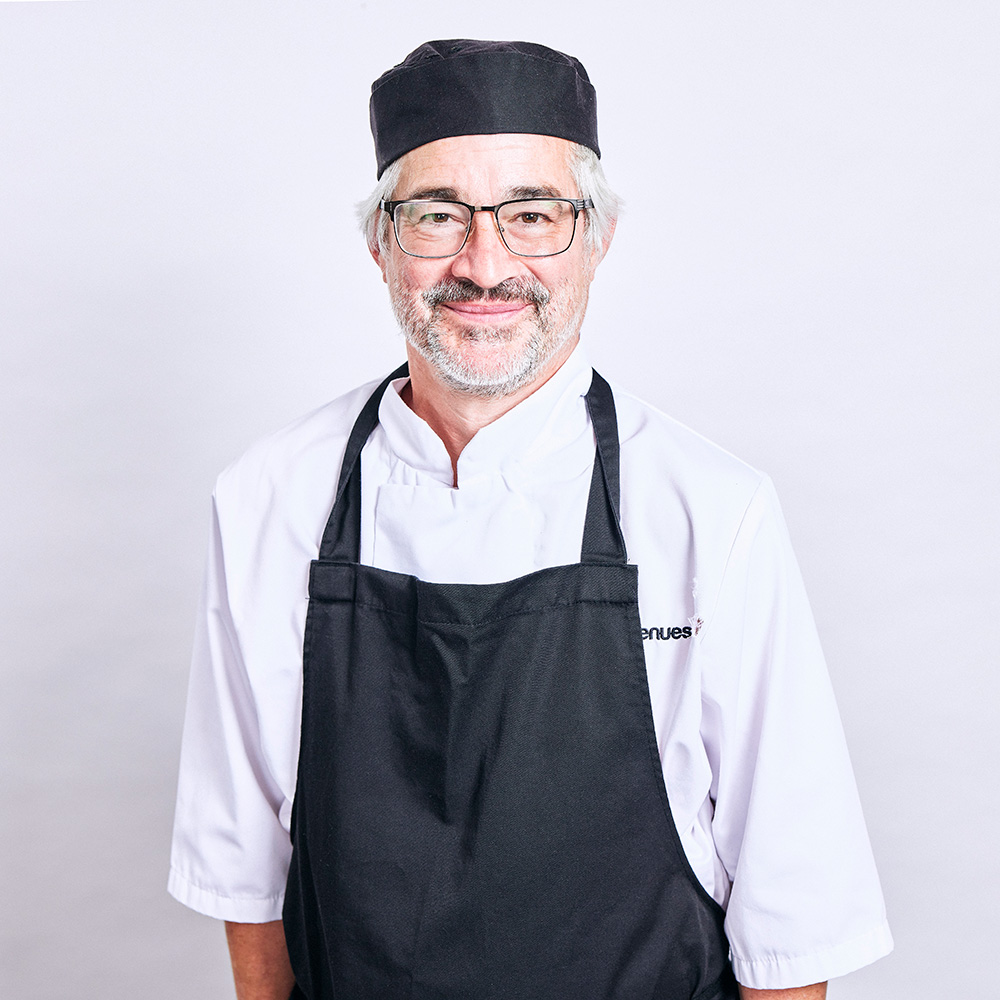 A headshot of a man with grey hair, a beard and glasses, wearing a chef's hat an apron, smiling at the camera.