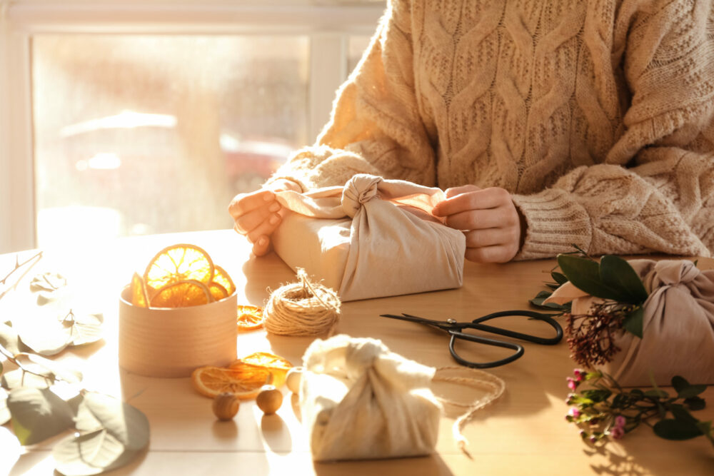 A person in a sweater wrapping a gift in fabric surrounded by craft decor on the table including dried flowers, vines, dried oranges and fabric.