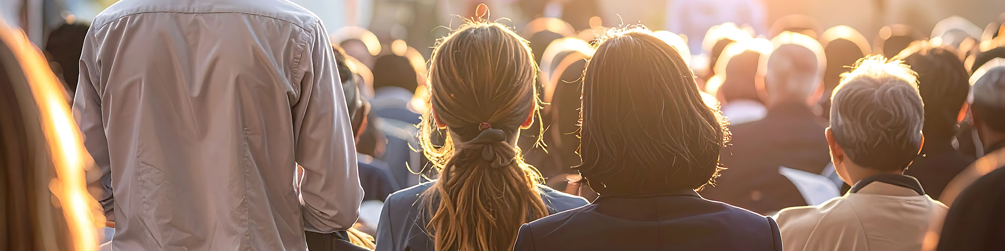 Behind people watching event talk outdoors with sun shining at camera.