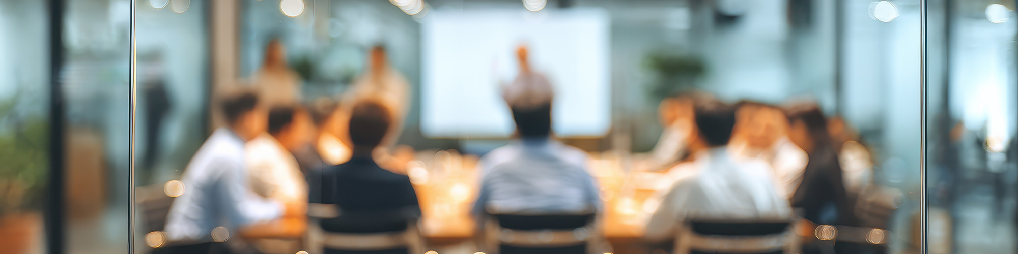Blurred image of people sitting in a meeting looking at screen.